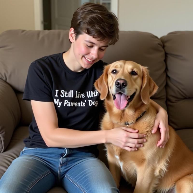 A young adult wearing an "I Still Live With My Parents' Dog" t-shirt, smiling and petting a Golden Retriever in a living room setting.